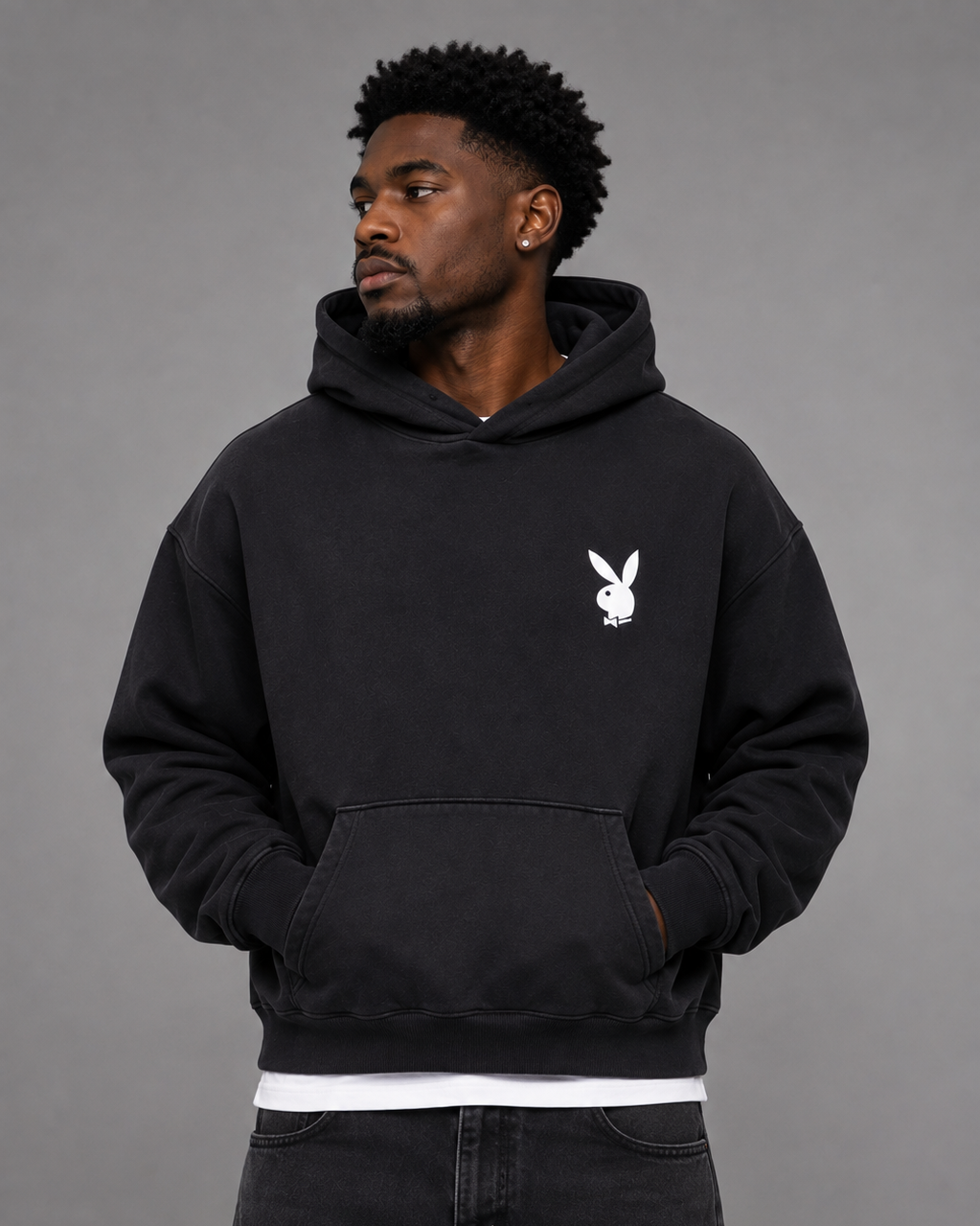 A man wearing a black pullover hoodie with a white Playboy bunny logo on the left chest, paired with black jeans and a white undershirt, against a solid gray studio background.