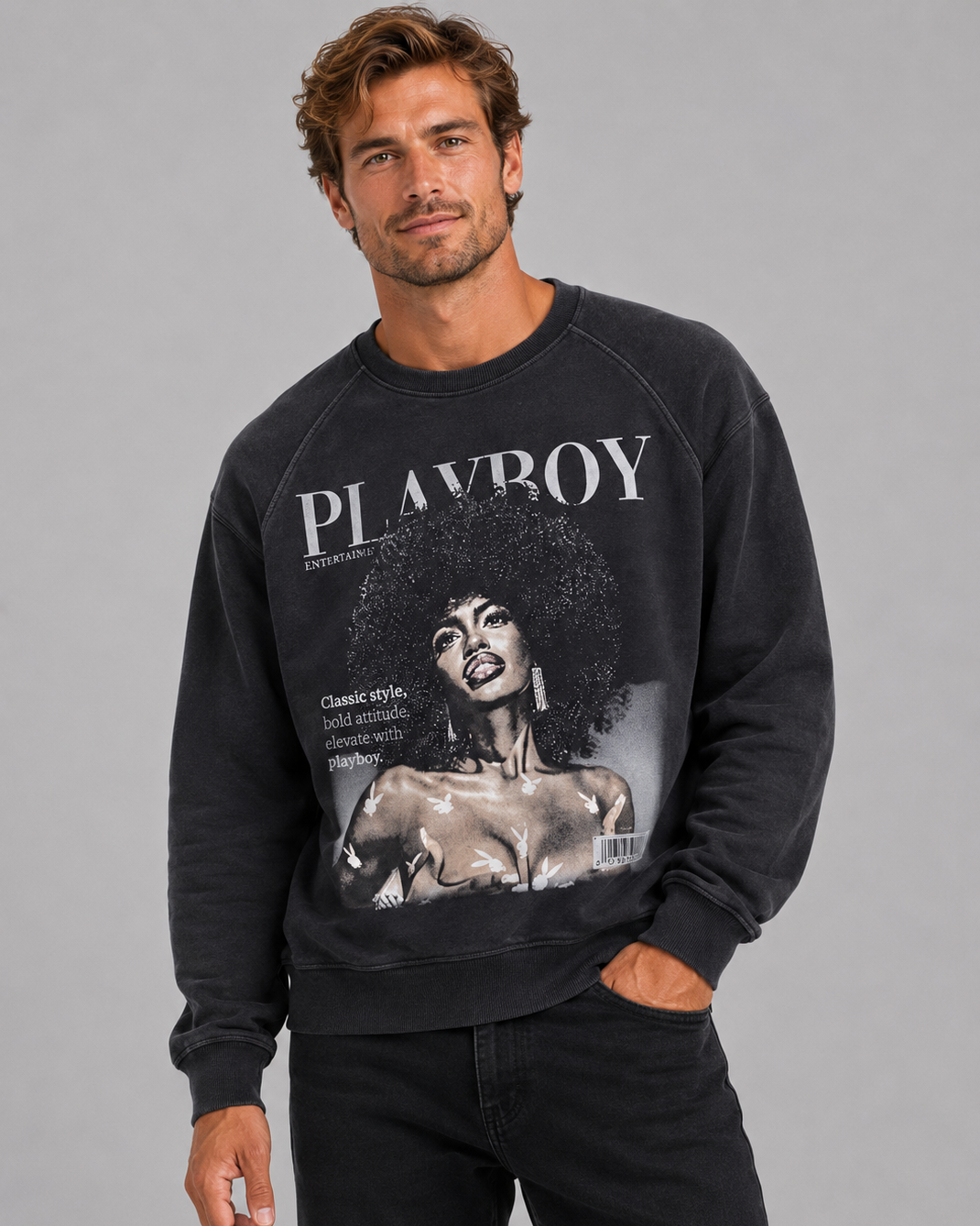 A man wearing a black Playboy sweatshirt with a large vintage-style graphic print on the front, paired with black jeans, against a solid gray studio background.