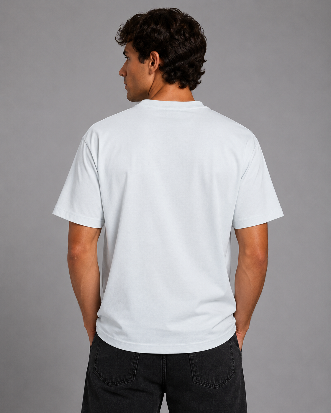 A man viewed from the back wearing a plain white short-sleeve crewneck t-shirt and dark jeans, standing against a solid gray studio background.