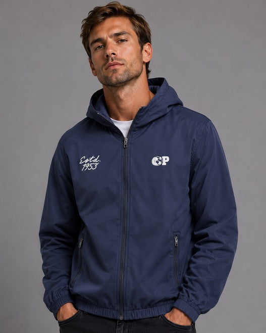 A man wearing a navy blue hooded zip-up jacket with white embroidered logos on the chest, paired with black jeans, standing against a solid gray studio background.