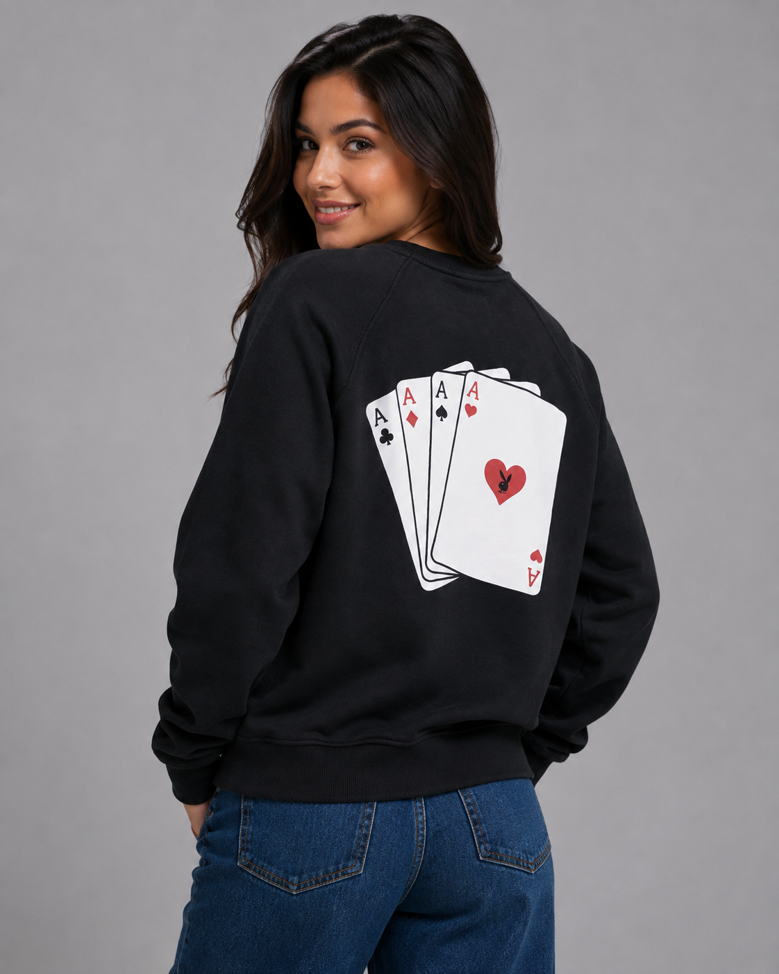 A woman wearing a black crewneck sweatshirt with a graphic of four aces of hearts on the back, paired with blue jeans, photographed against a solid gray studio background. #Women