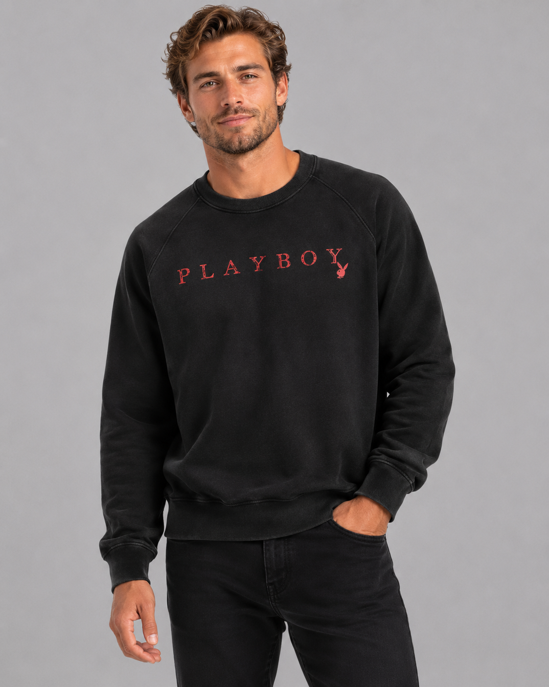 A man wearing a black crewneck sweatshirt with 'PLAYBOY' printed in red across the chest, paired with black jeans, against a solid gray studio background.