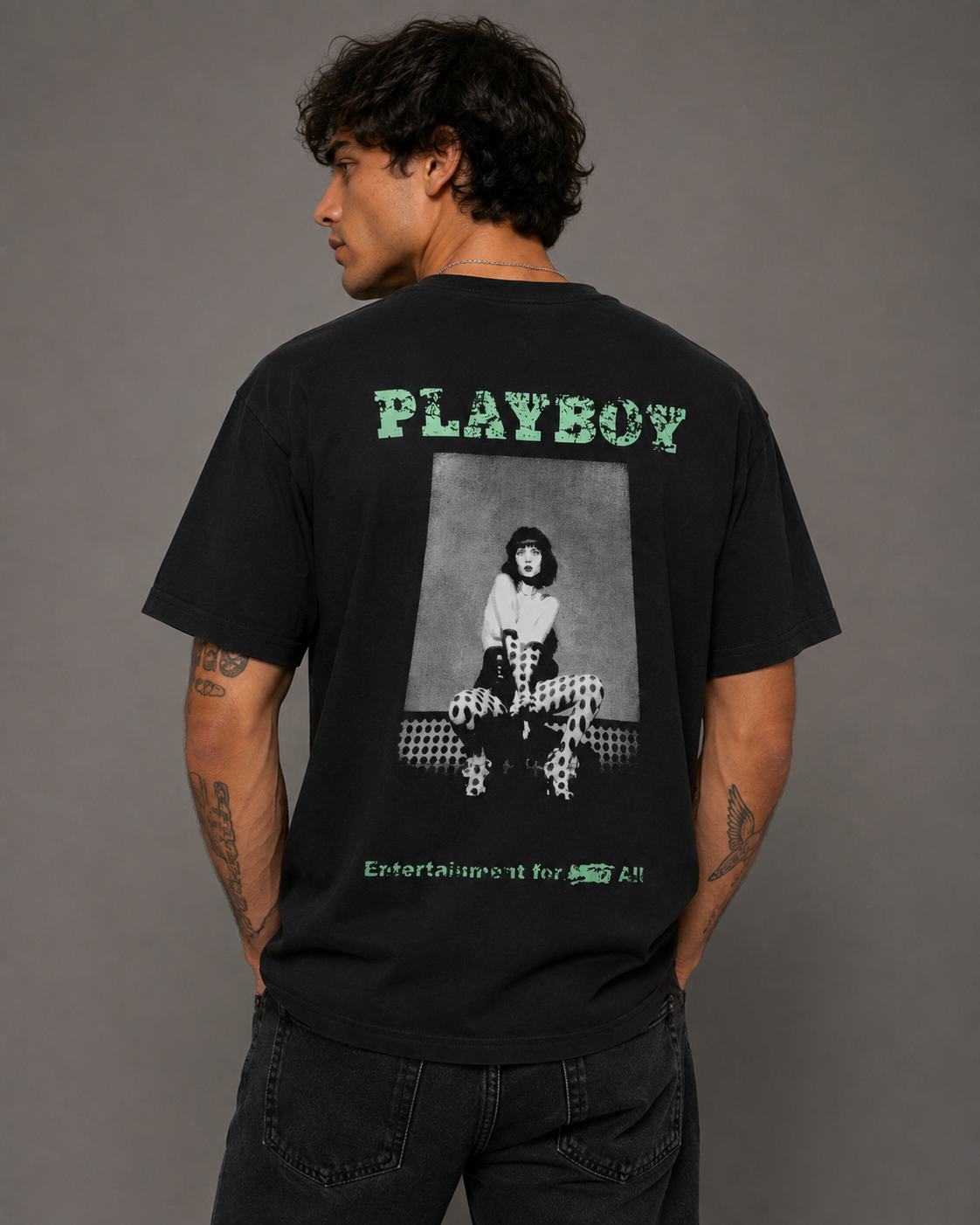 A man wearing a black Playboy t-shirt, viewed from the back. The shirt features a large graphic print on the back with the brand name 'PLAYBOY' in green above a black-and-white photo of a woman, and green text below reading 'Entertainment for All'.