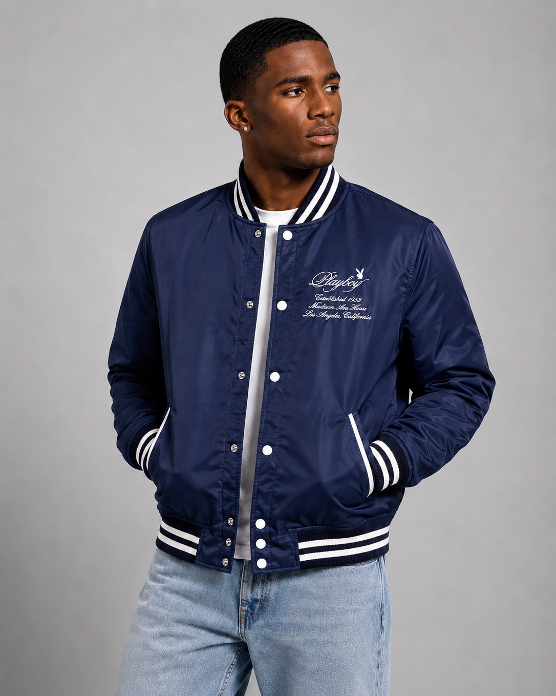 A man wearing a navy blue varsity bomber jacket with white stripes and embroidered script on the chest, paired with light blue jeans, against a solid gray studio background.