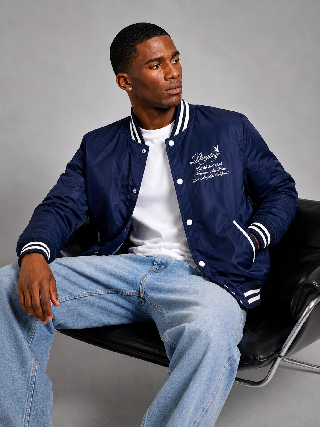 A man wearing a navy blue varsity jacket with white stripes and embroidered script on the chest, styled over a white t-shirt and light blue jeans, sitting on a black leather chair against a gray studio background.