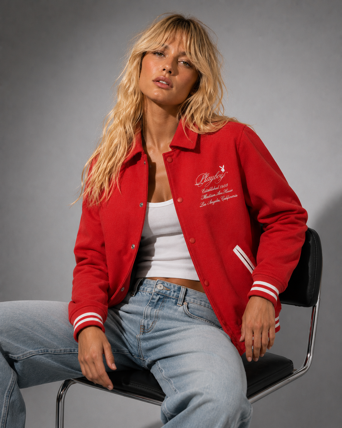 A blonde woman sits on a black chair wearing a bright red varsity-style jacket with white embroidered script on the chest and white striped ribbed cuffs, paired with a white tank top and light blue jeans against a gray studio background. #Women