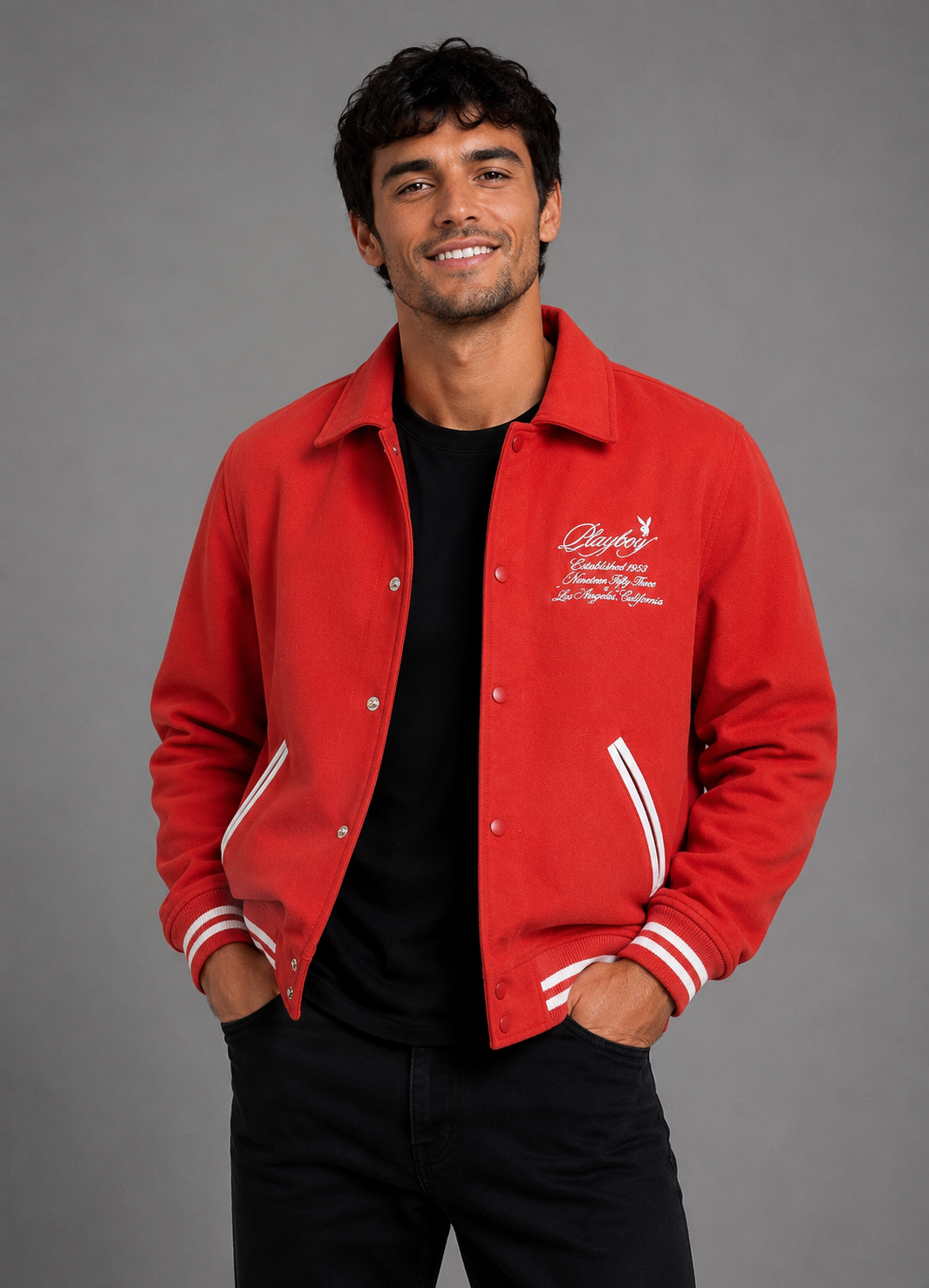 A man wearing a bright red varsity-style jacket with white embroidered script on the left chest and white striped ribbed cuffs and hem, layered over a black t-shirt and black pants, against a solid gray studio background.