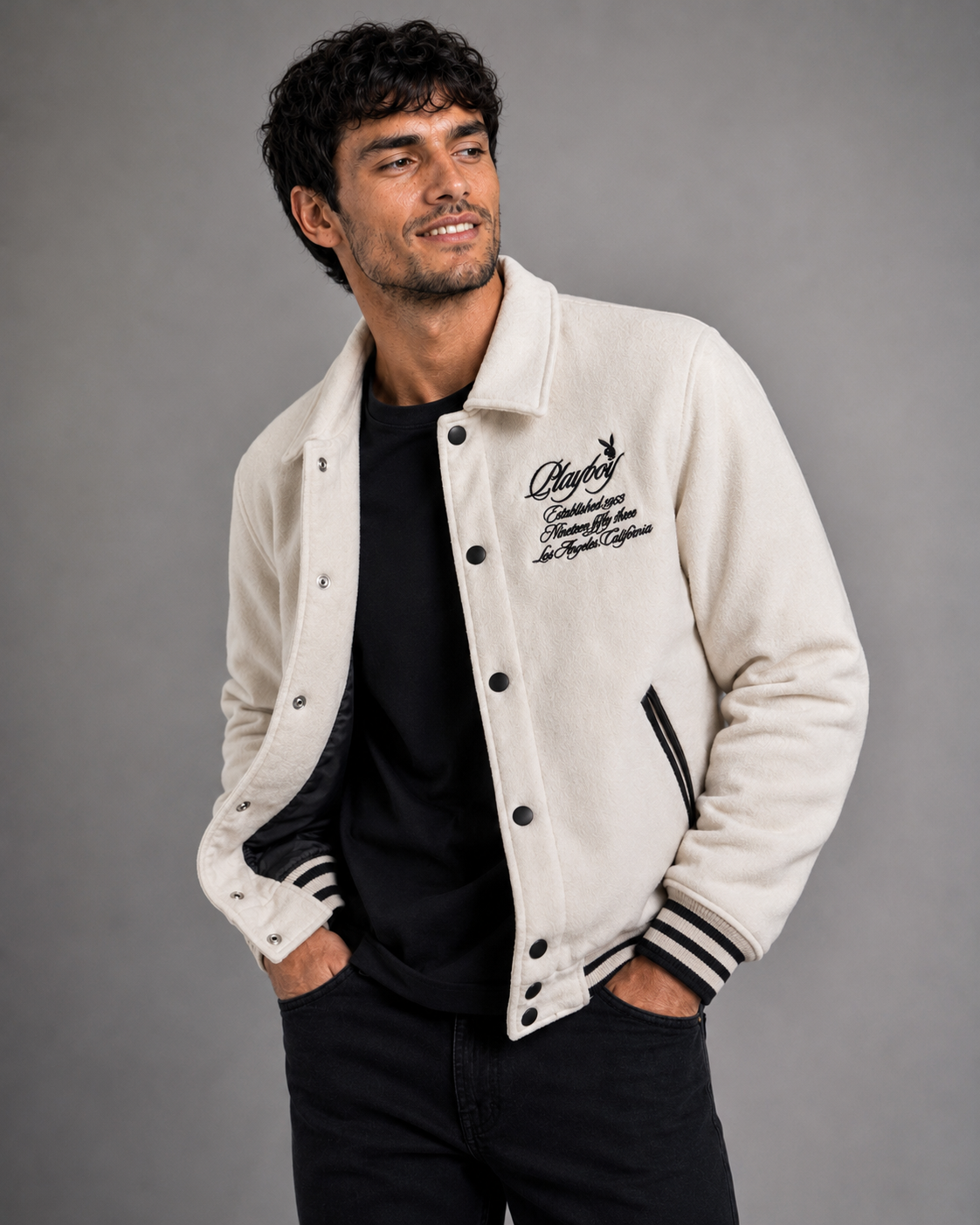 A man wearing a cream-colored varsity jacket with black buttons, black and white striped ribbed cuffs and hem, and black embroidered script on the left chest, paired with a black t-shirt and black jeans, against a solid gray studio background.