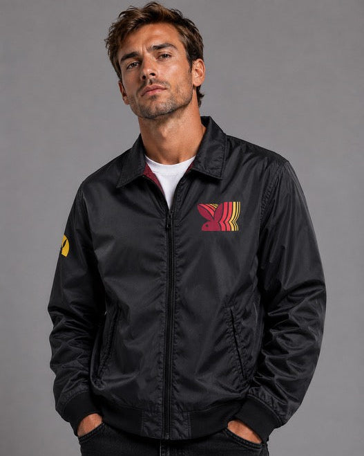 A man wearing a black nylon bomber jacket with a red and yellow chest logo, photographed against a solid gray studio background.