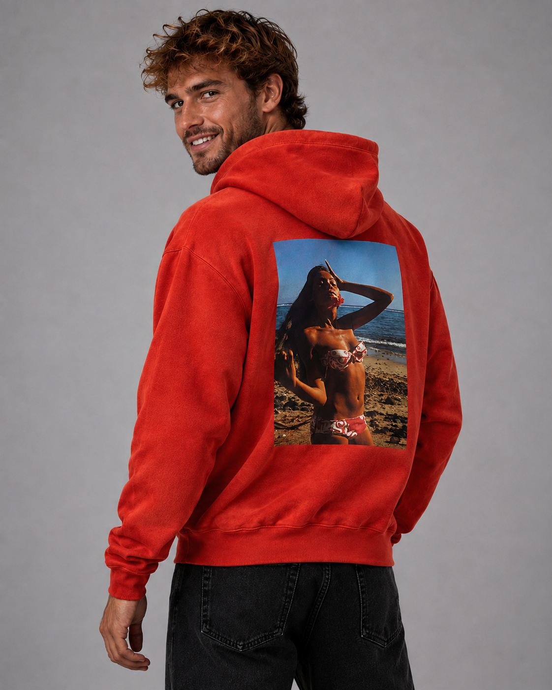 A man wearing a bright red hoodie with a large rectangular beach photo print on the back, photographed against a plain gray studio background.