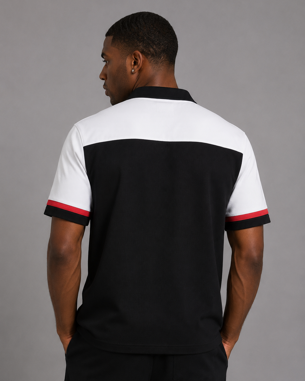 A man viewed from the back wearing a color-blocked short-sleeve polo shirt with a black body, white yoke, and red and white sleeve trim, paired with black trousers, against a solid gray studio background.
