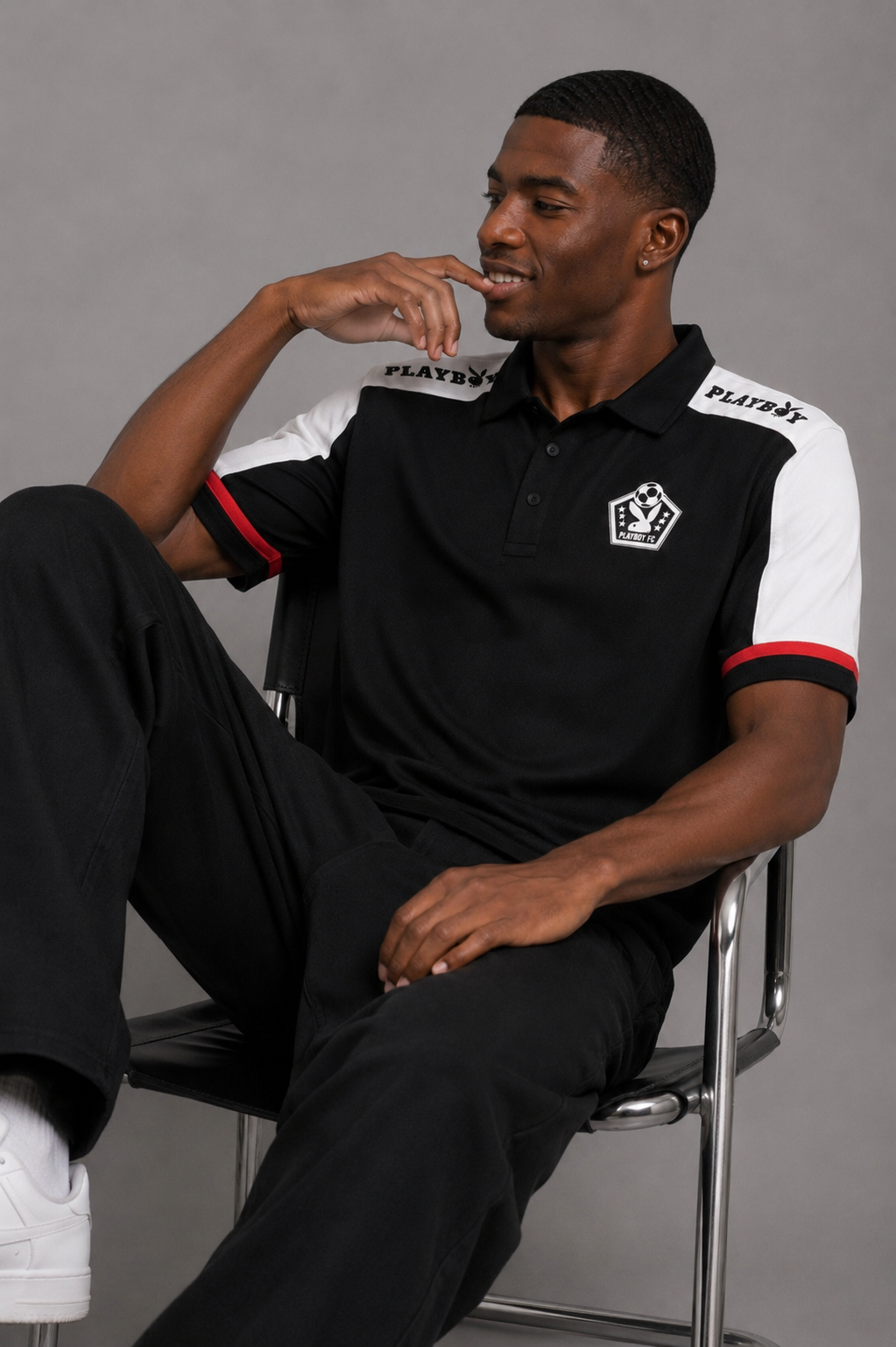 A man wearing a black and white short-sleeve polo shirt with red accents and the brand name 'PLAYBOY' on the shoulders, sitting on a metal chair against a gray studio background.