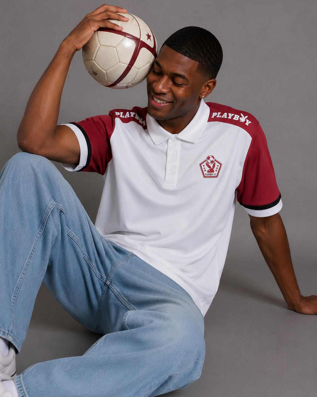 A man wearing a white and maroon short-sleeve polo shirt with 'PLAYBOY' printed on the shoulders, light blue jeans, and holding a soccer ball against his head, posed against a solid gray studio background.