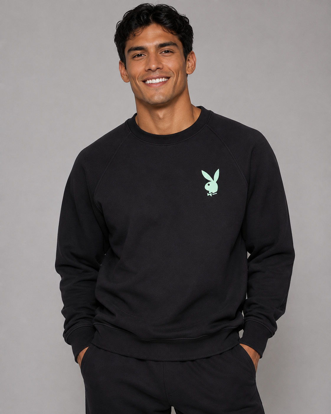 A smiling man wearing a black crewneck sweatshirt with a small mint green Playboy bunny logo on the left chest, paired with black sweatpants, against a solid gray studio background.