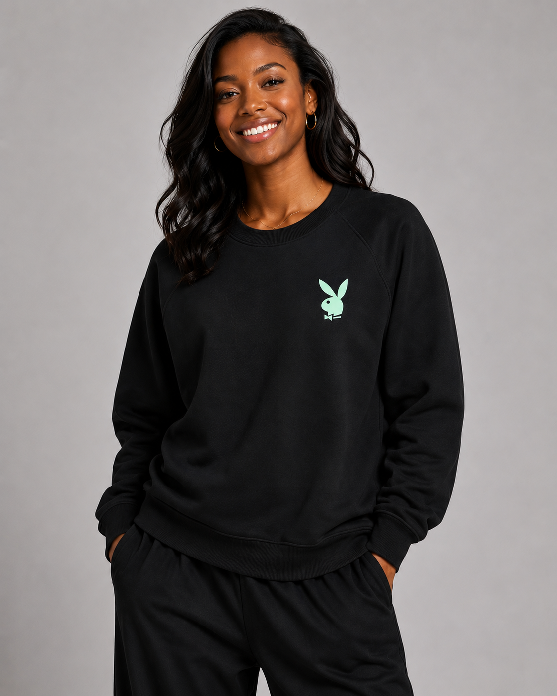 A woman wearing a black crewneck sweatshirt with a small mint green Playboy bunny logo on the left chest, paired with black sweatpants, against a solid gray studio background. #Women