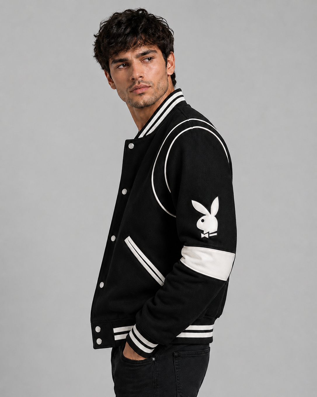 A man wearing a black varsity-style jacket with white stripes and a white Playboy bunny logo on the sleeve, posed against a solid gray studio background.