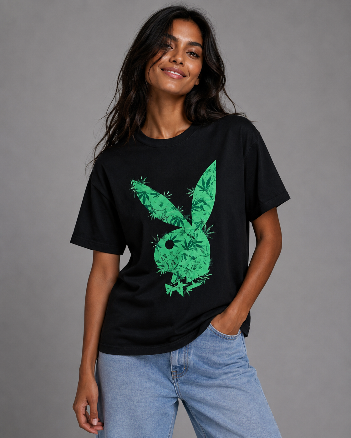 A woman wearing a black short-sleeve t-shirt with a green Playboy bunny graphic filled with leaf patterns, paired with light blue jeans, against a solid gray studio background. #Women