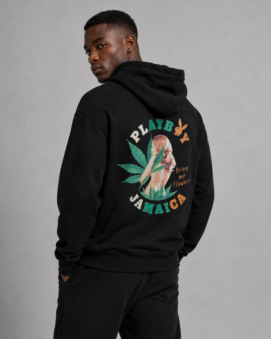 A man wearing a black hoodie with a large back graphic that reads 'PLAYBOY JAMAICA' and features a colorful illustration, photographed against a neutral gray studio background.