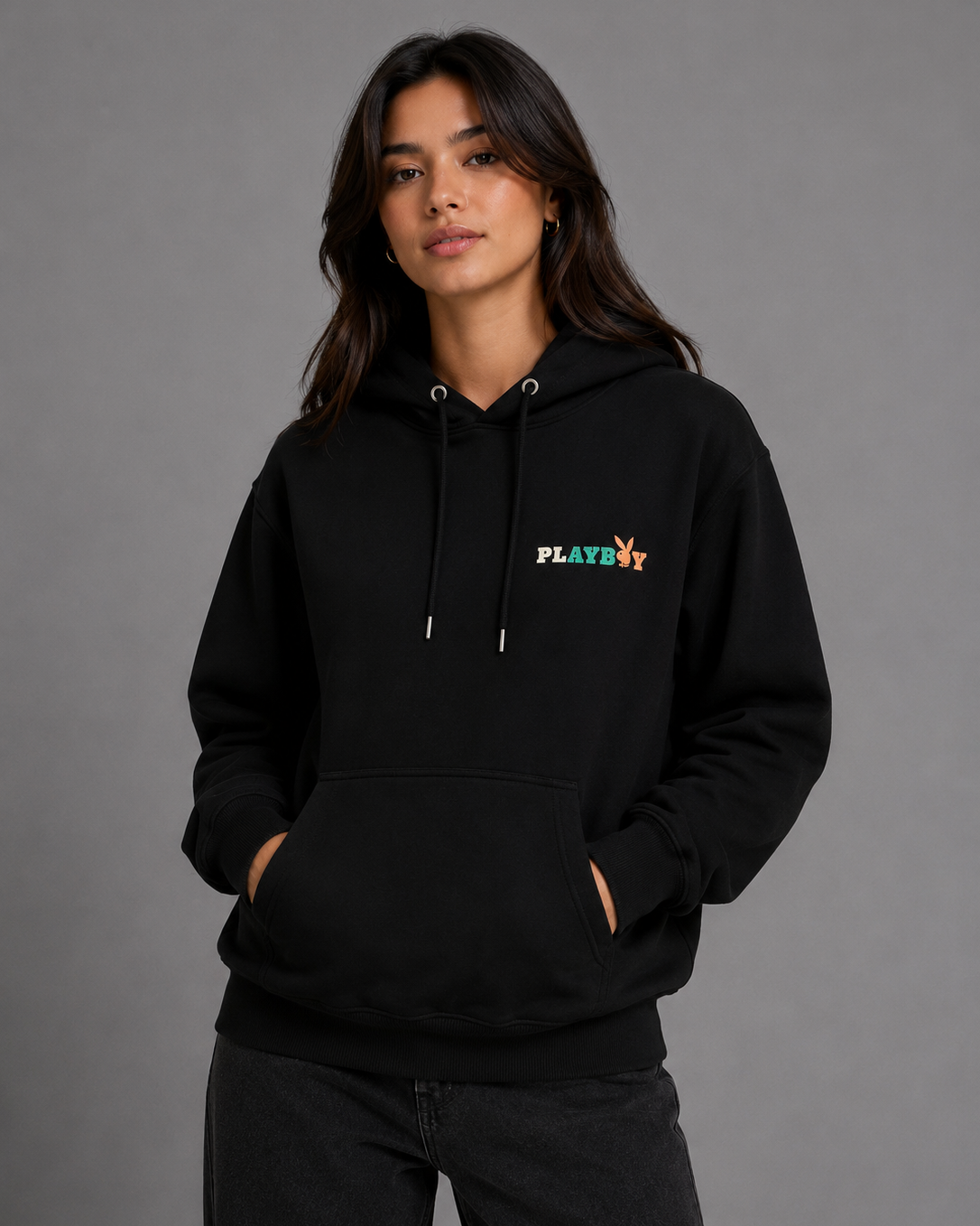 A woman wearing a black pullover hoodie with a small multicolor PLAYBOY logo on the left chest, posed against a solid gray studio background. #Women