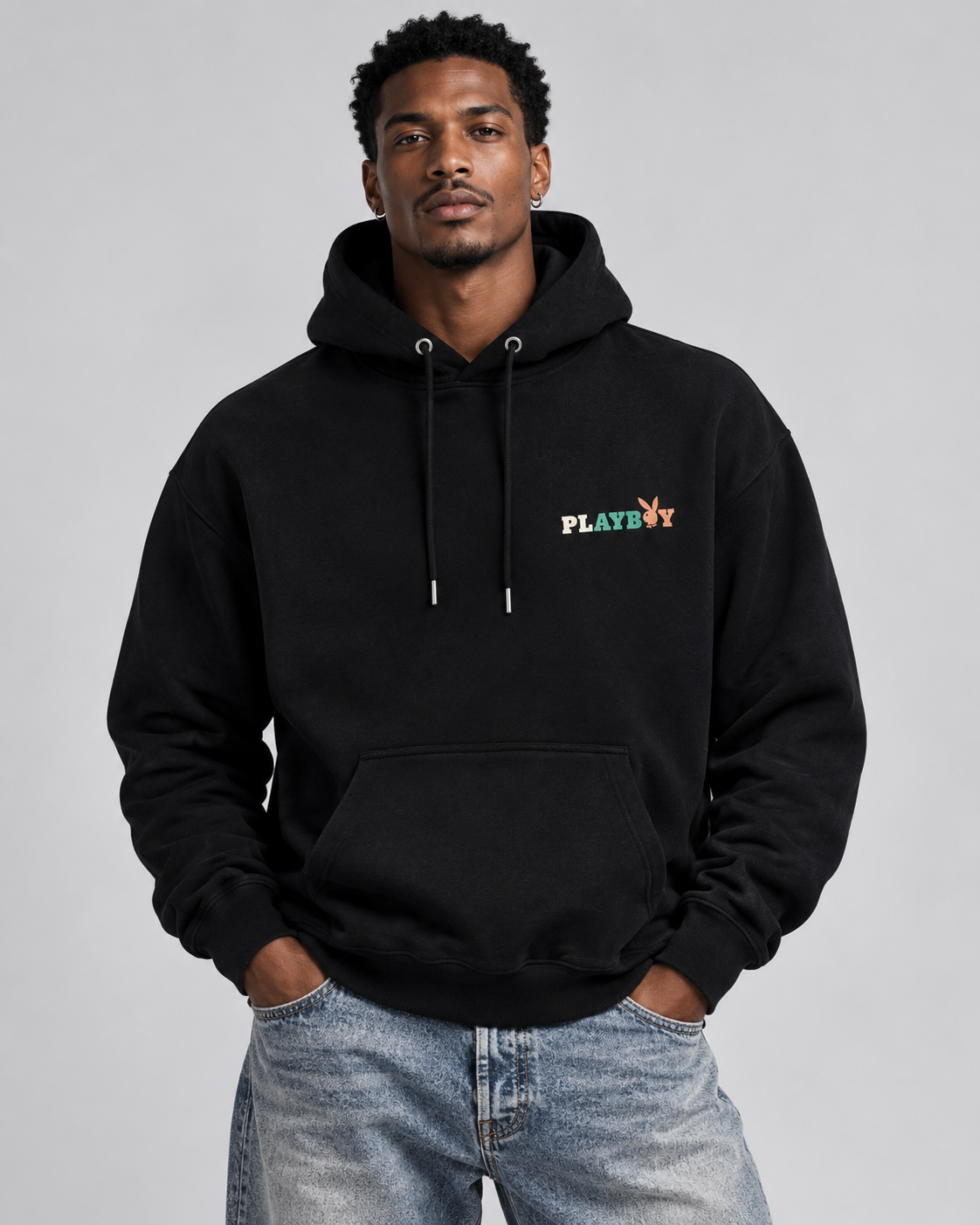 A man wearing a black pullover hoodie with a small multicolor PLAYBOY logo on the chest, paired with light-wash blue jeans, against a solid gray studio background.