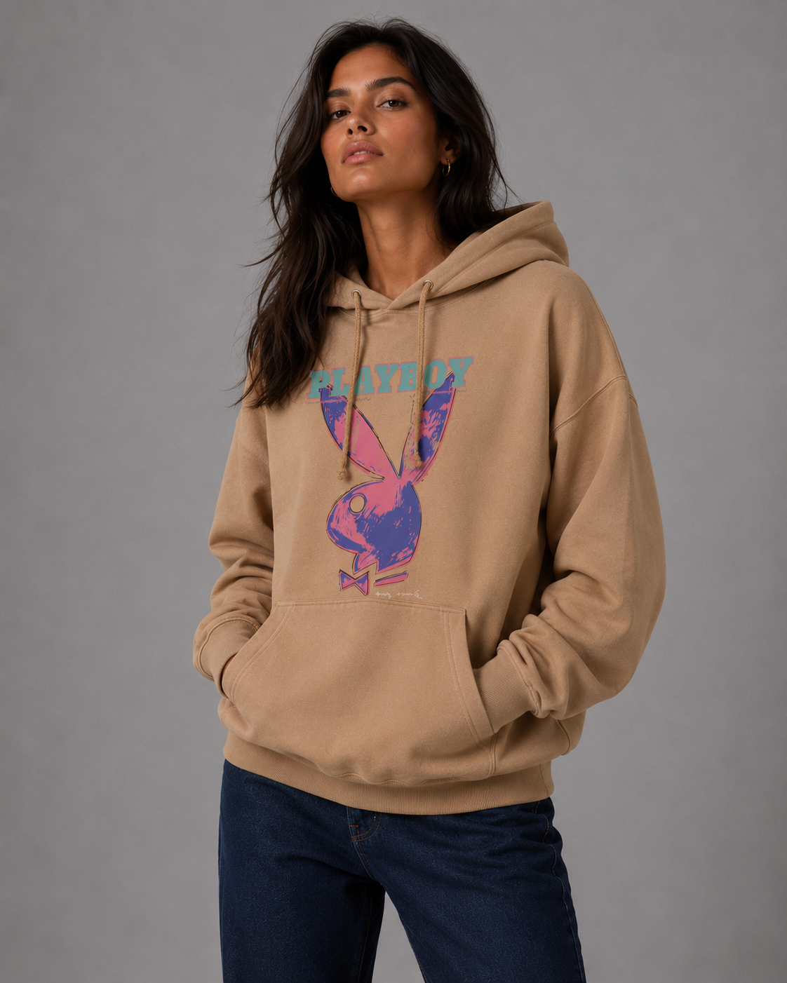 A woman wearing a tan hoodie with a colorful Playboy bunny graphic and the word 'PLAYBOY' across the chest, paired with dark blue jeans, against a solid gray studio background. #Women