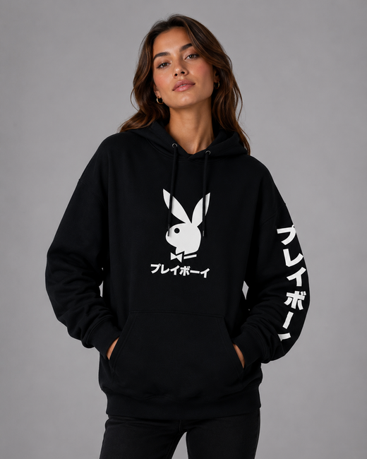 A woman modeling a black pullover hoodie with a white Playboy bunny logo and Japanese text on the front, plus white Japanese text down one sleeve, against a plain gray studio background.