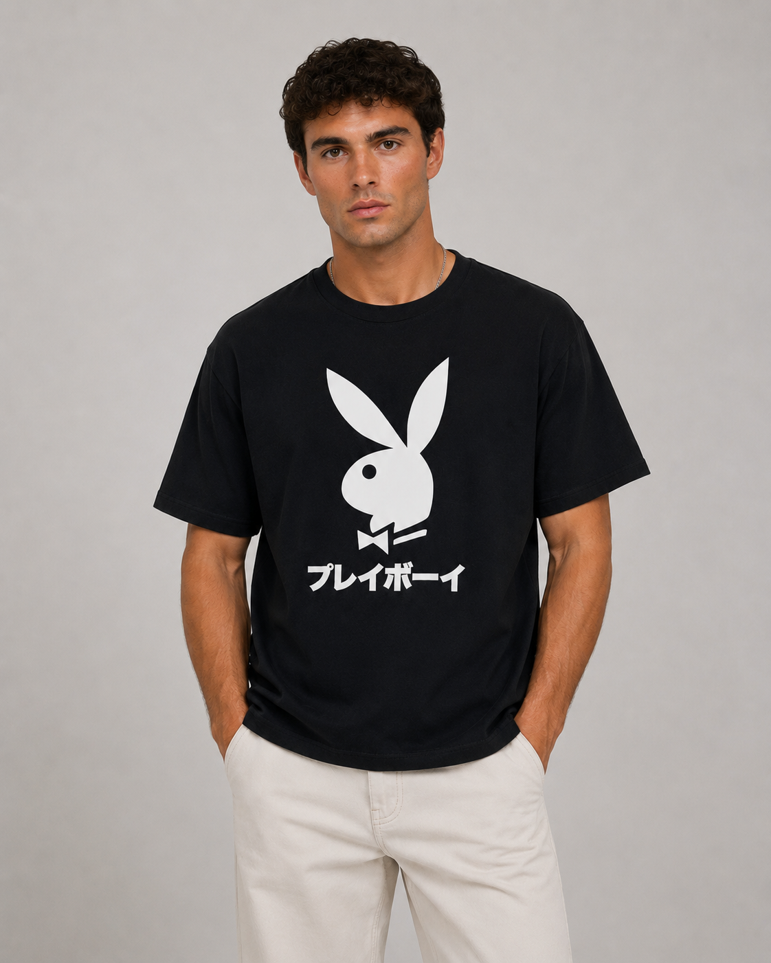 A man wearing a black short-sleeve t-shirt with a large white Playboy bunny logo and Japanese katakana text on the front, paired with off-white pants, against a plain gray studio background.