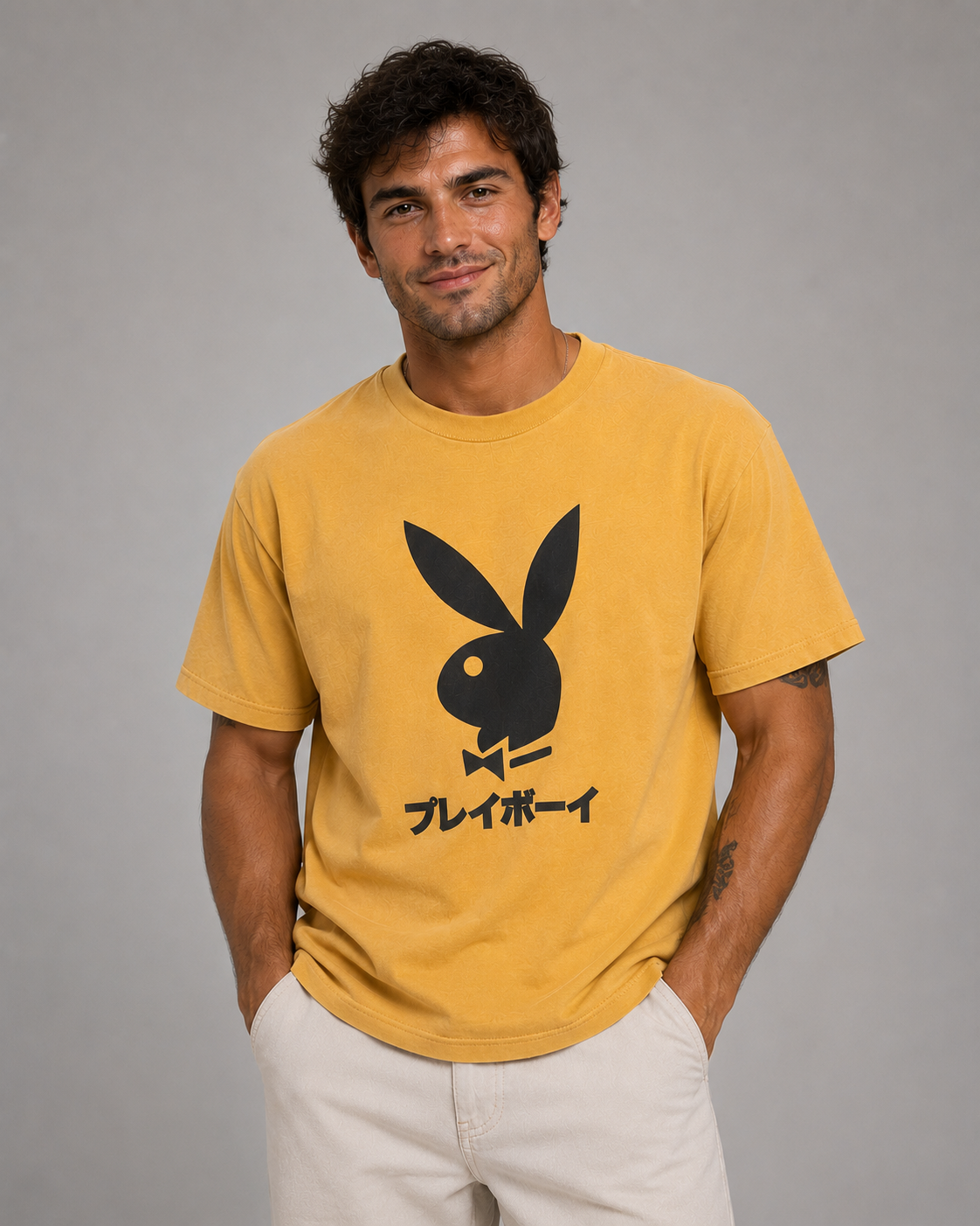 A man wearing a yellow short-sleeve t-shirt with a black Playboy bunny logo and Japanese text on the front, paired with off-white pants, against a solid gray studio background.