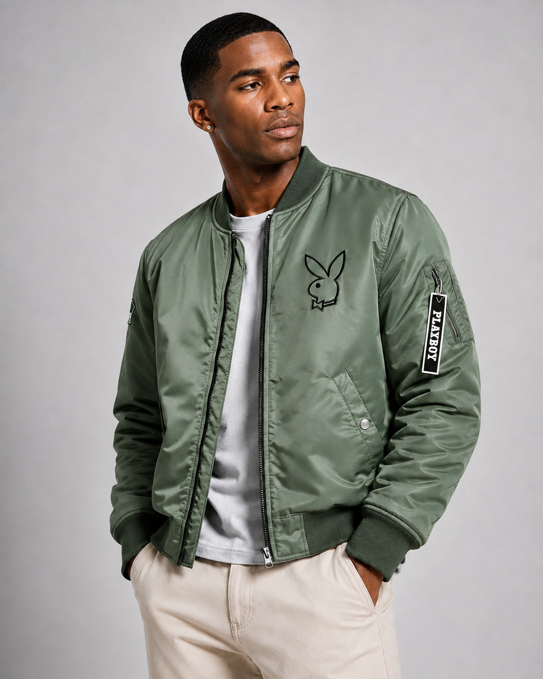A man wearing an olive green bomber jacket with a black Playboy bunny logo on the left chest and a sleeve patch, styled over a white t-shirt and beige pants against a gray studio background.