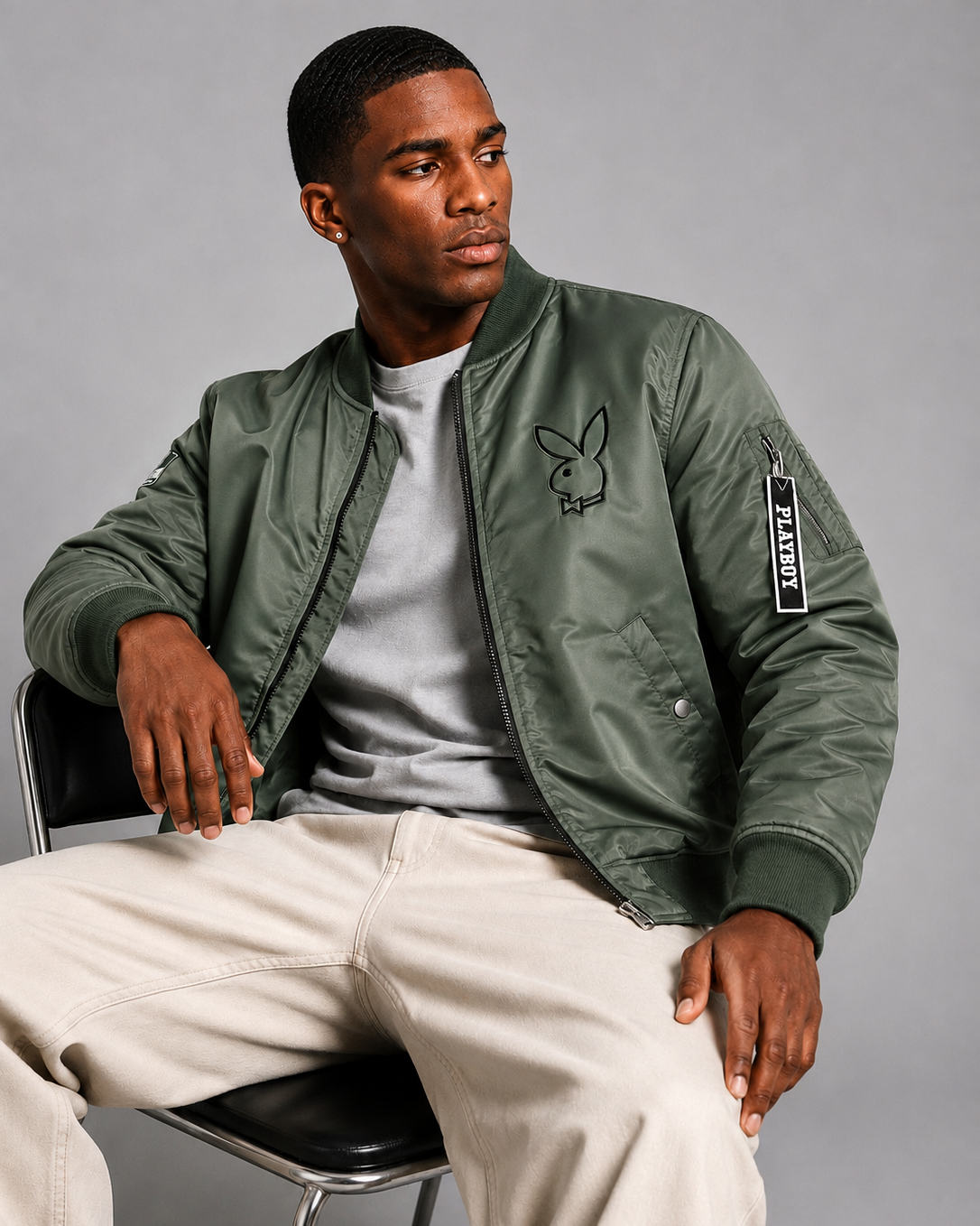 A man wearing an olive green bomber jacket with a black Playboy bunny logo on the chest and a vertical Playboy sleeve patch, paired with a gray t-shirt and beige pants, sitting on a black chair against a gray studio background.