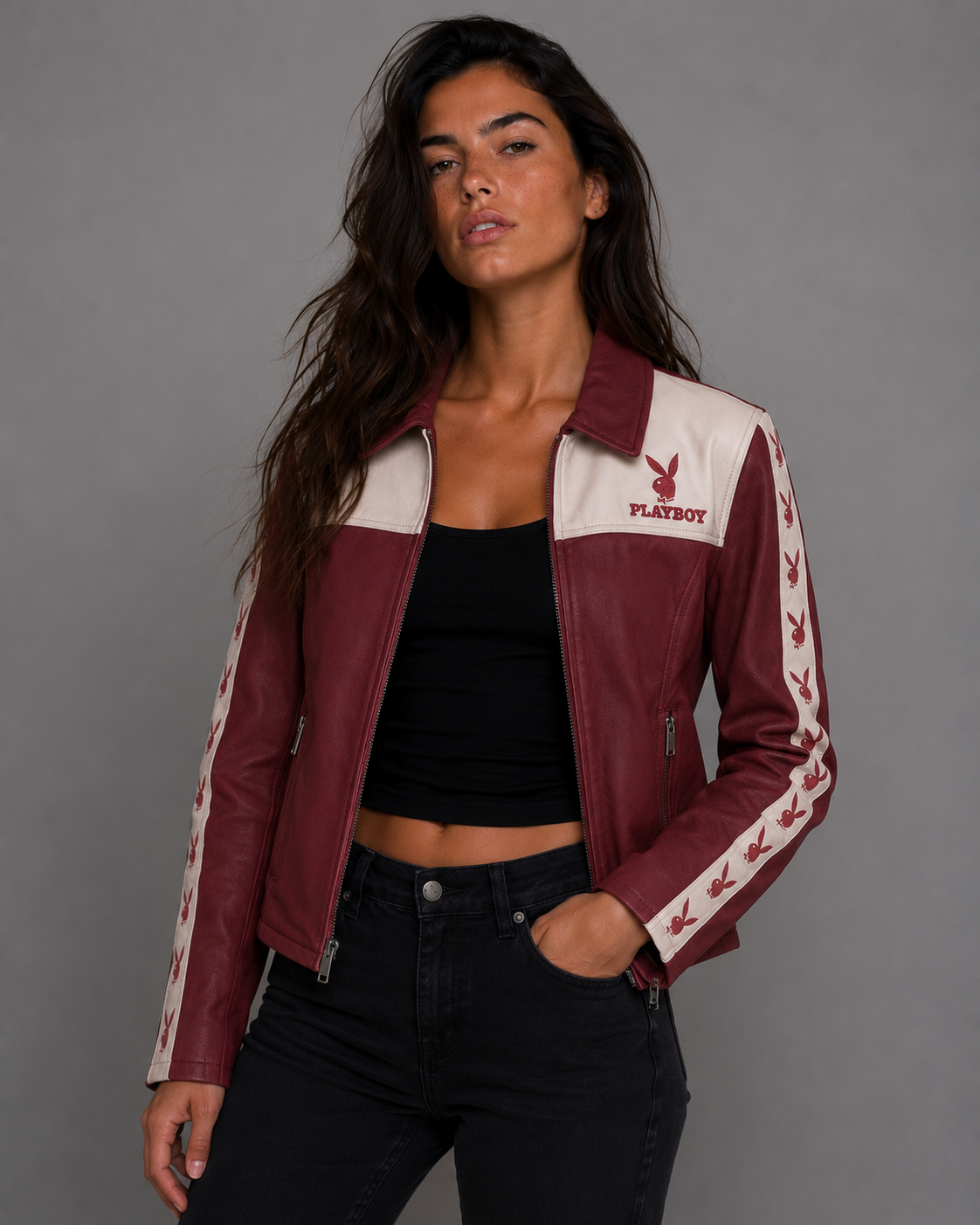 A woman wearing a maroon and cream color-blocked leather jacket with Playboy branding on the chest and sleeves, paired with a black crop top and black jeans, photographed against a neutral gray studio background. #Women