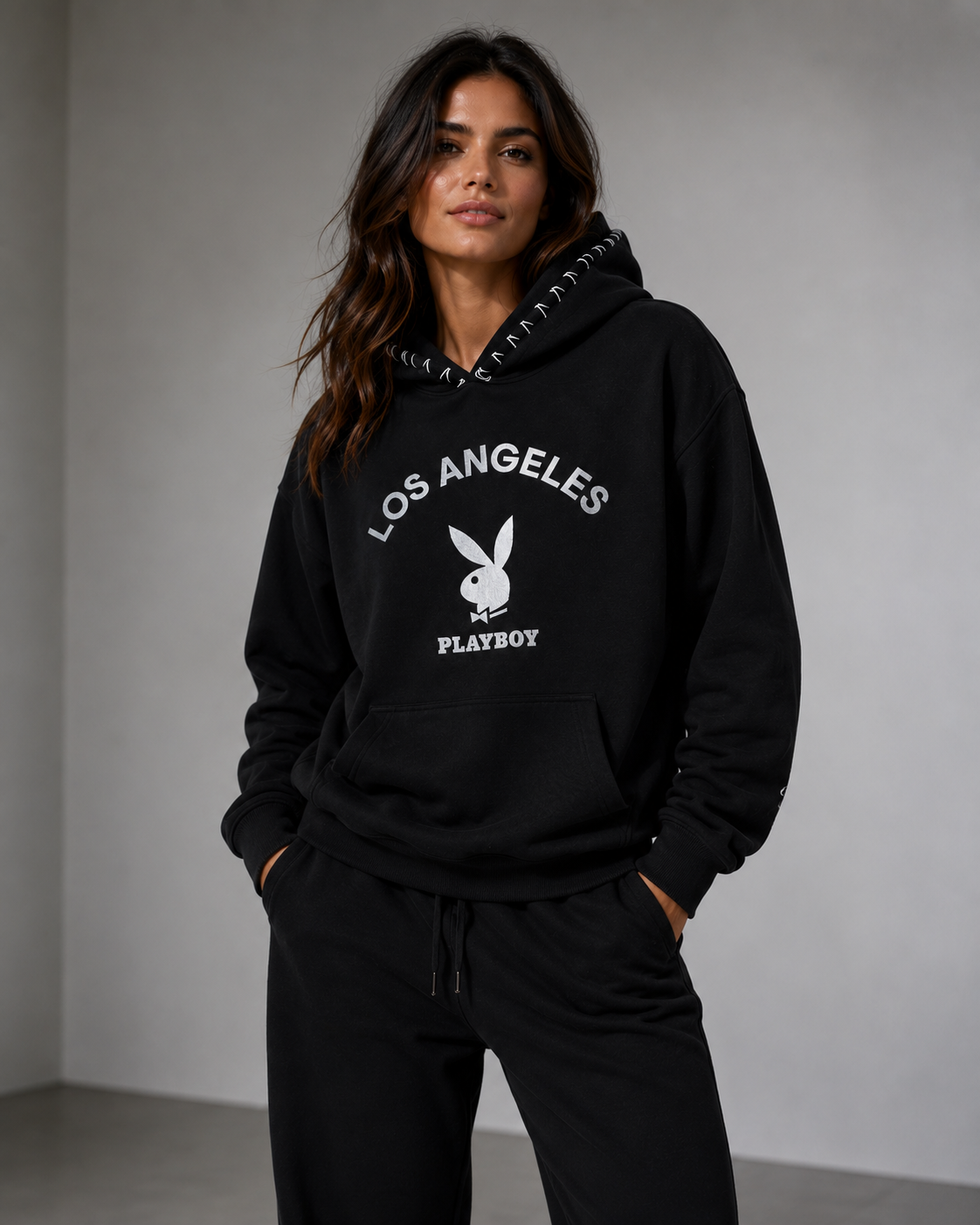 A woman wearing a black Los Angeles Playboy hoodie with white graphic text and logo on the front, paired with black sweatpants, standing against a neutral gray wall. #Women