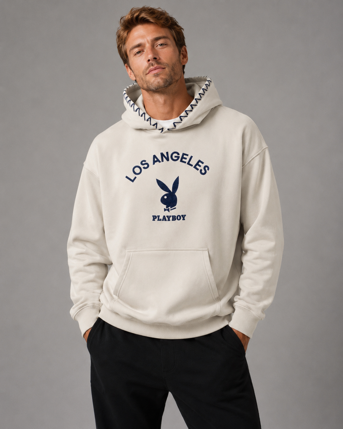 A man wearing a cream-colored Playboy hoodie with a navy blue 'Los Angeles' graphic and bunny logo on the front, paired with black pants, against a solid gray studio background.