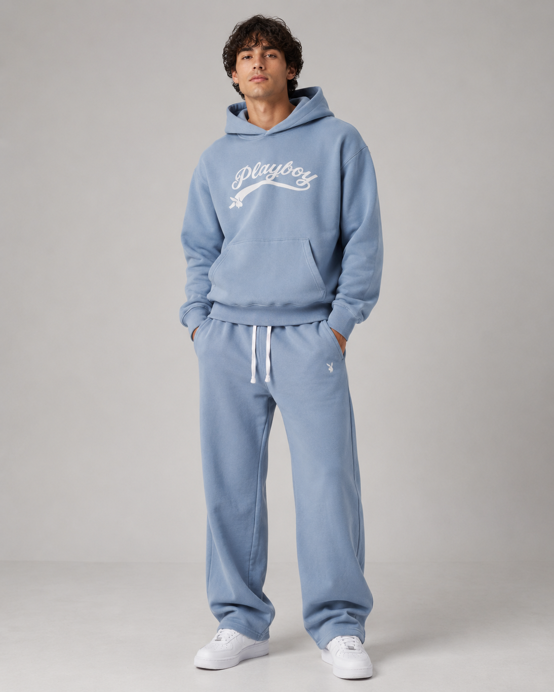 A man modeling a light blue matching hoodie and sweatpants set with white drawstrings and white sneakers, standing against a neutral gray studio background.