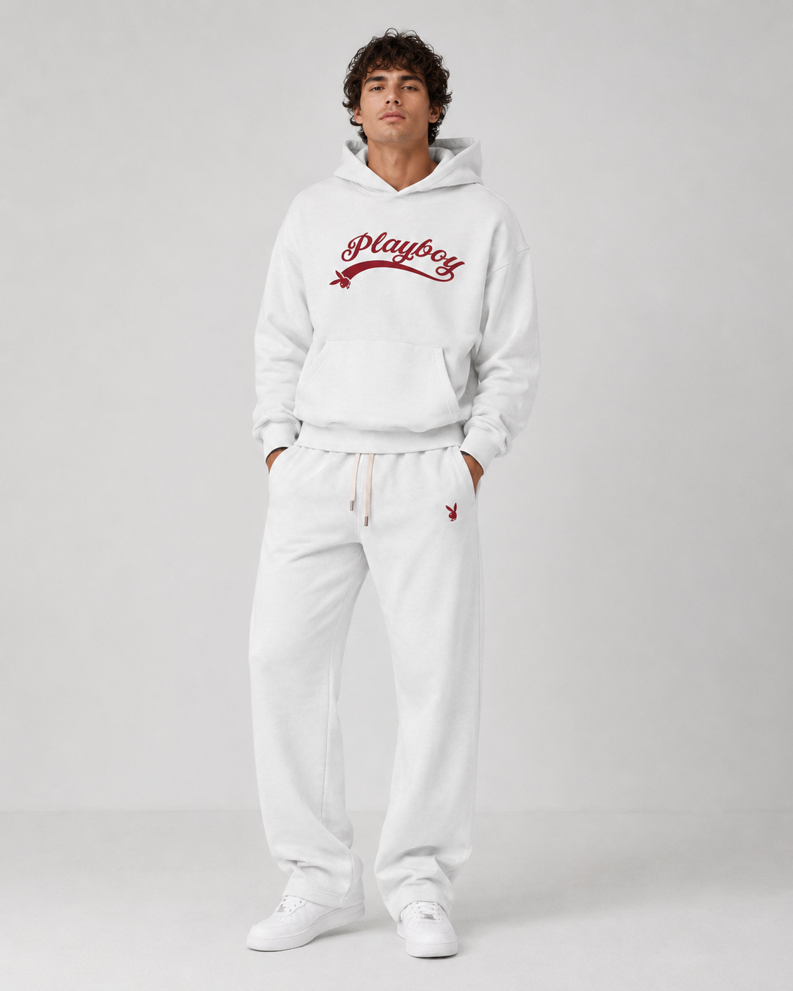 A man modeling a white sweatshirt and sweatpants set from Playboy, featuring a red script logo on the chest and a small red logo on the thigh, set against a plain light gray studio background.