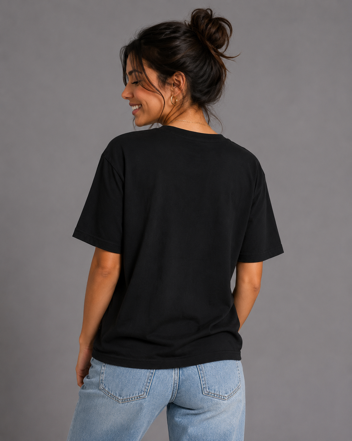 A woman with her hair in a bun is shown from the back wearing a plain black short-sleeve t-shirt and light blue jeans against a solid gray studio background. #Women