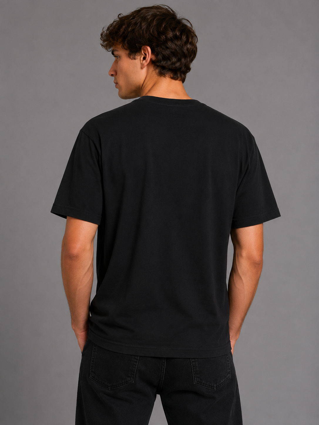 A man viewed from the back wearing a plain black short-sleeve crewneck t-shirt and black jeans, standing against a solid gray studio background.