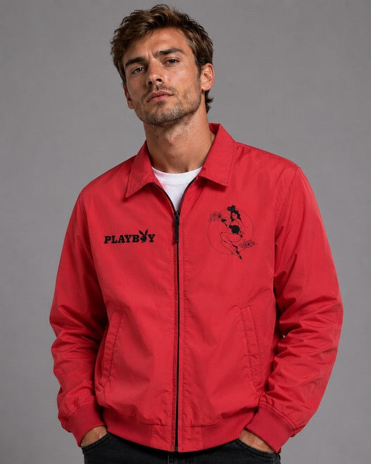 A man wearing a bright red zip-up jacket with black embroidered graphics, including the word 'PLAYBOY' on the left chest and a horse logo on the right chest, over a white t-shirt and dark jeans, against a solid gray studio background.