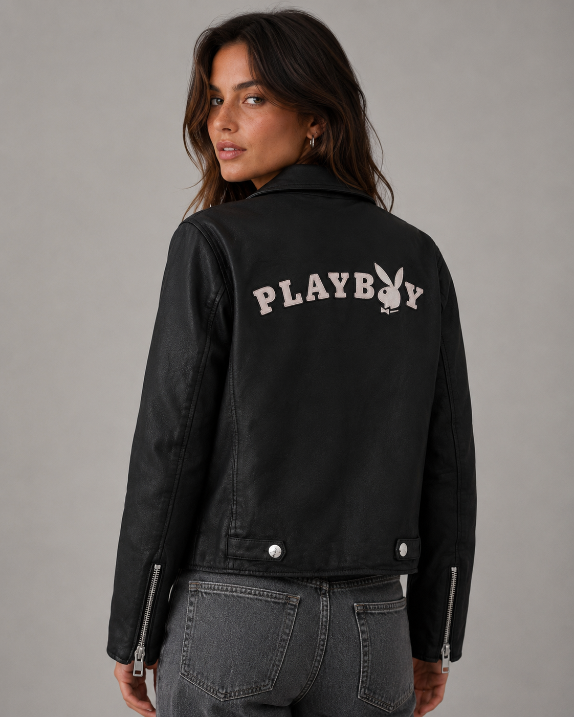 A woman wearing a black leather jacket with 'PLAYBOY' and the bunny logo embroidered in white on the back, paired with gray jeans, photographed against a neutral gray studio background. #Women