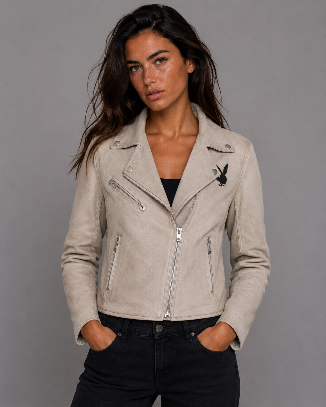 A woman wearing a light beige faux-leather moto jacket with silver zippers and a black Playboy bunny logo on the chest, paired with black jeans, against a solid gray studio background. #Women