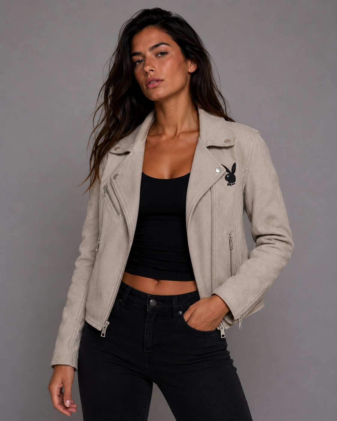 A woman modeling a light beige faux-suede moto jacket with a black embroidered bunny patch on the chest, worn over a black crop top and black jeans against a gray studio background. #Women