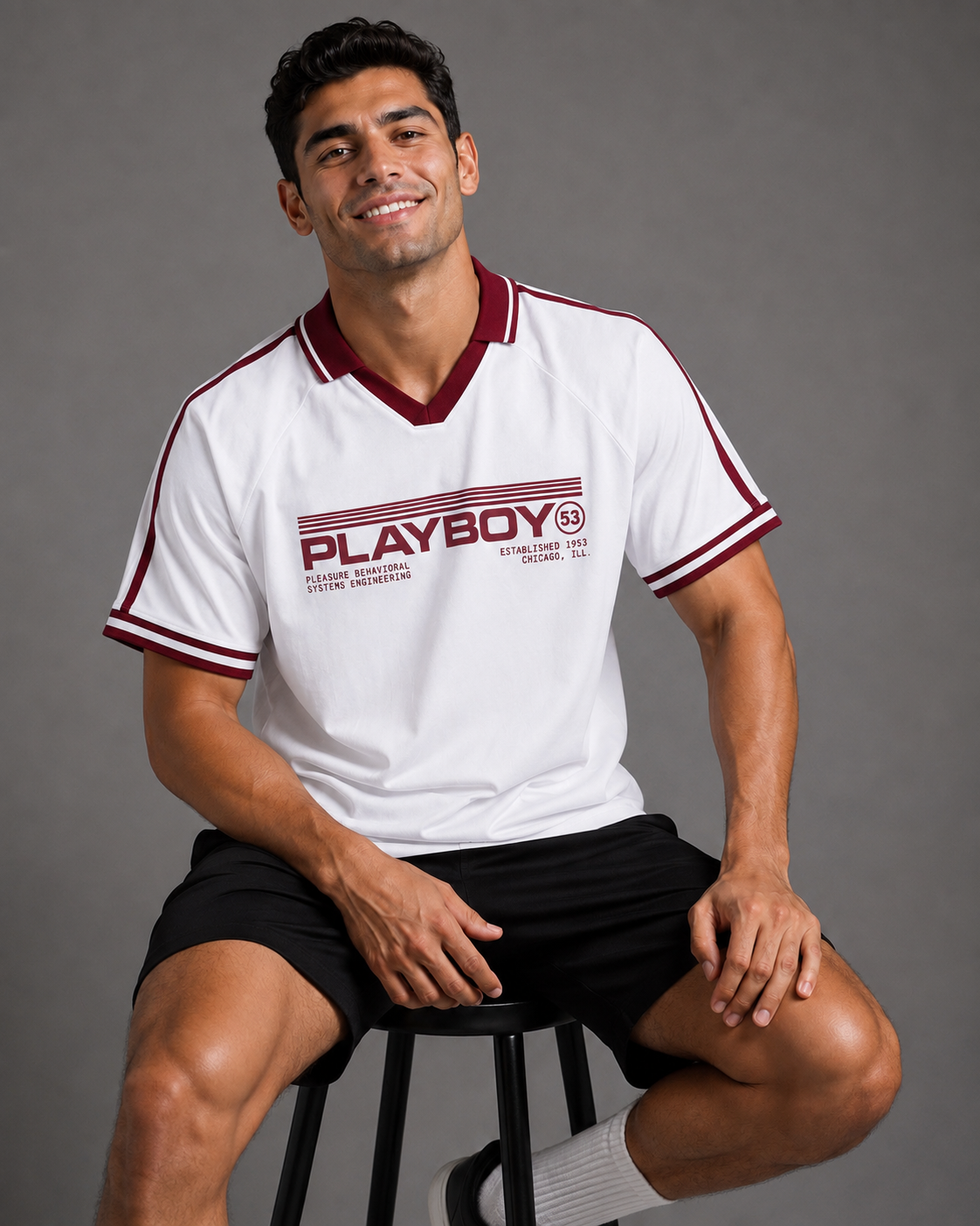 A man wearing a white Playboy short-sleeve V-neck t-shirt with maroon trim and black shorts, sitting on a stool against a gray studio background.