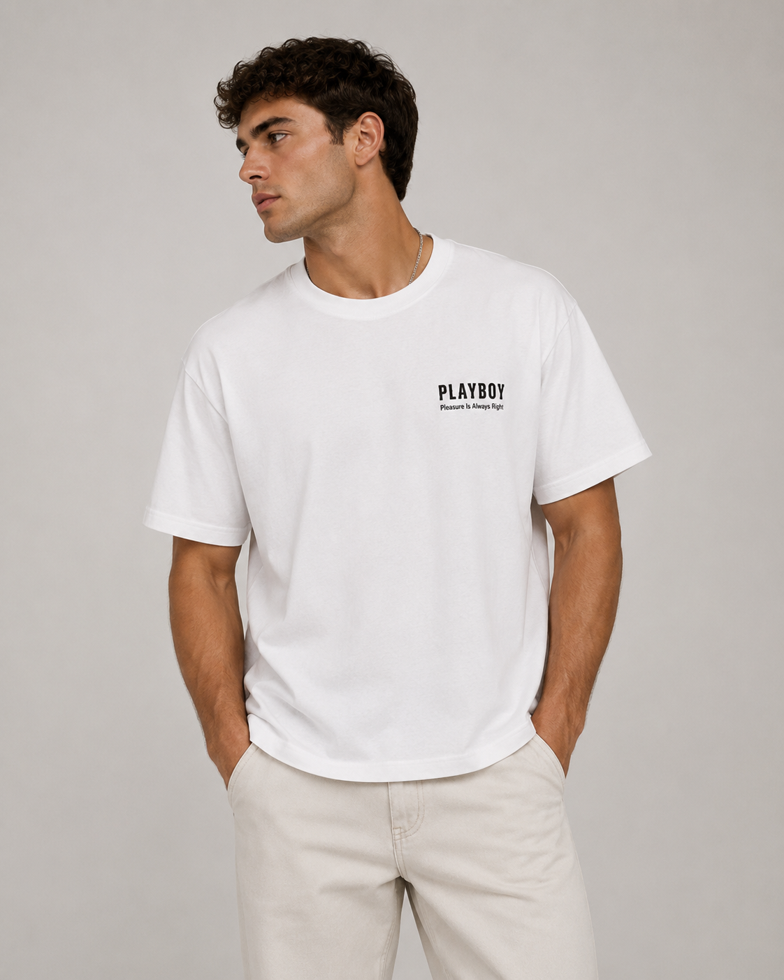A man wearing a white short-sleeve crewneck t-shirt with a black PLAYBOY logo on the left chest, paired with light beige trousers, against a solid light gray background.