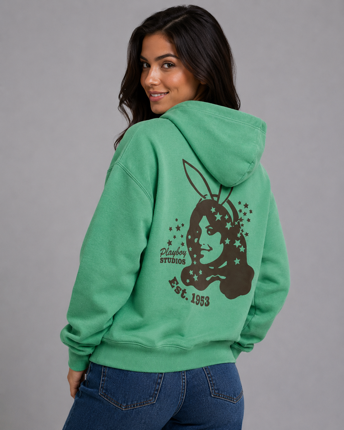 A woman wearing a green hoodie with a dark brown Playboy bunny graphic on the back, paired with blue jeans, photographed against a solid gray studio background. #Women