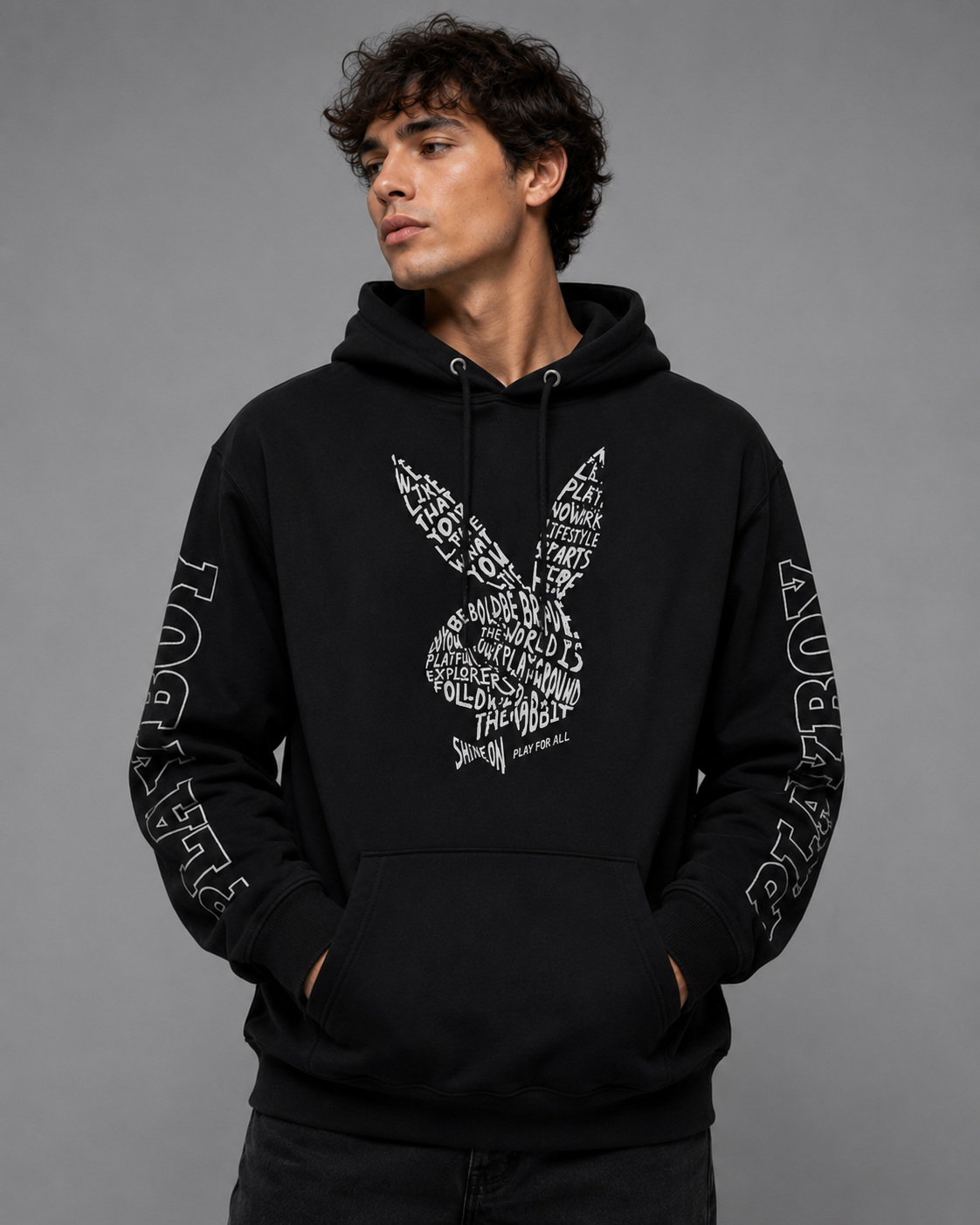 A man wearing a black pullover hoodie with a white typographic graphic on the front and 'PLAYBOY' text down both sleeves, photographed against a solid gray studio background.