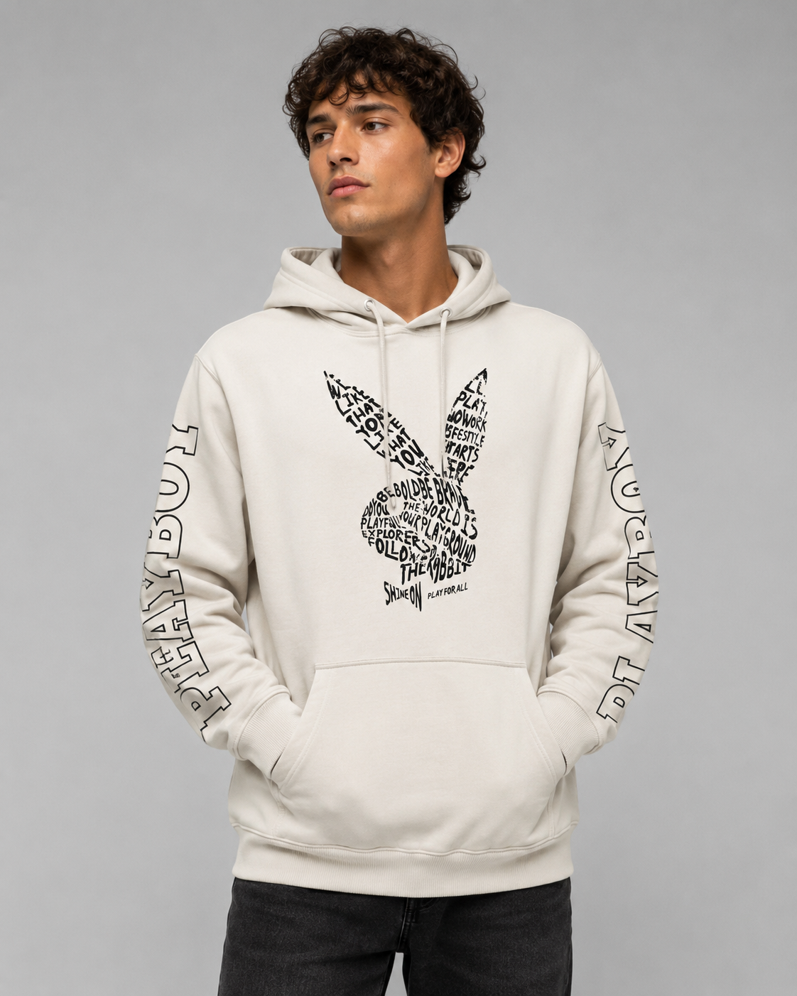 A man wearing a light beige Playboy hoodie with a black graphic print on the front and 'PLAYBOY' text down both sleeves, posed against a solid gray studio background.