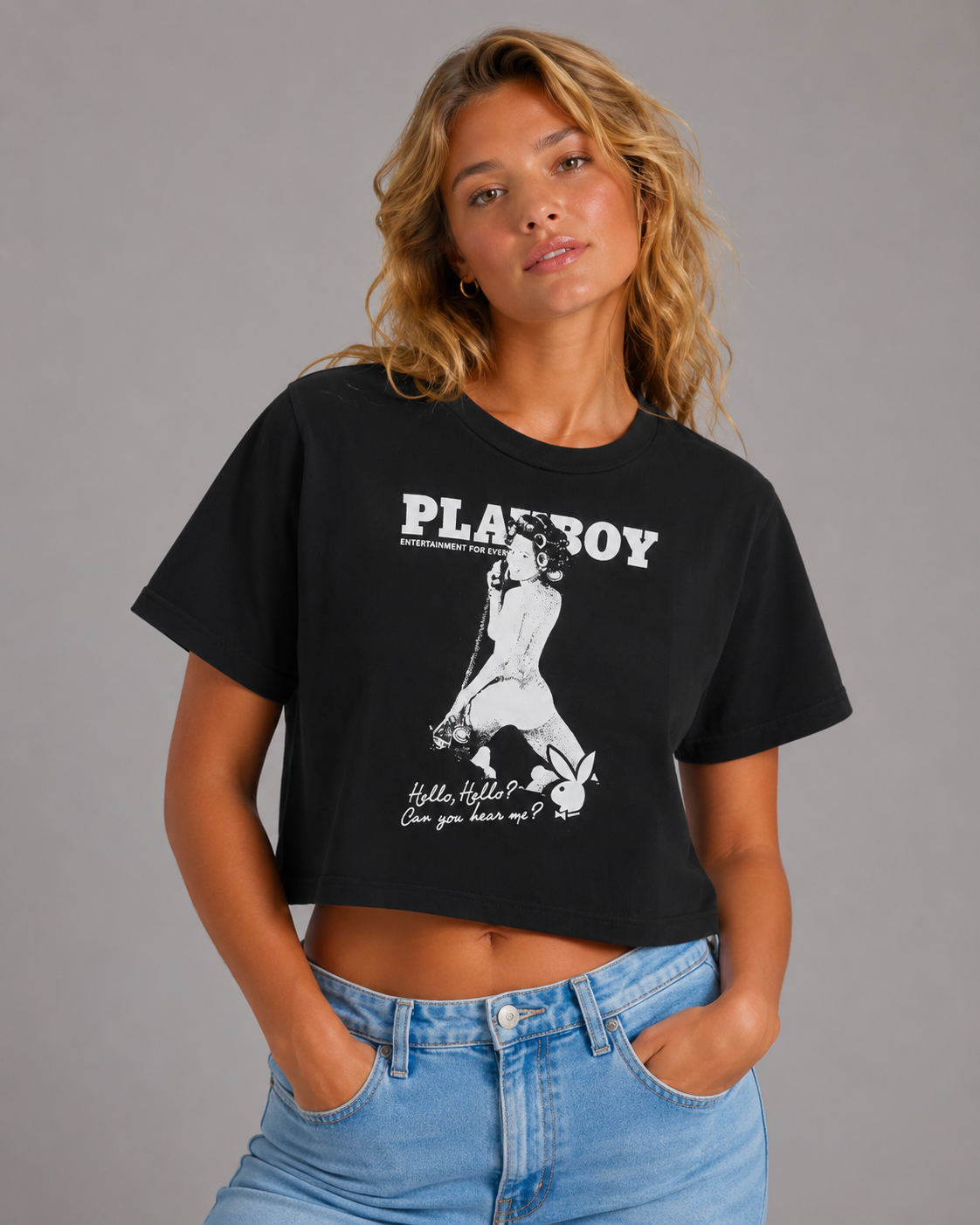 A woman wearing a black cropped graphic t-shirt with a white Playboy logo and illustration, paired with light blue jeans, against a solid gray studio background. #Women