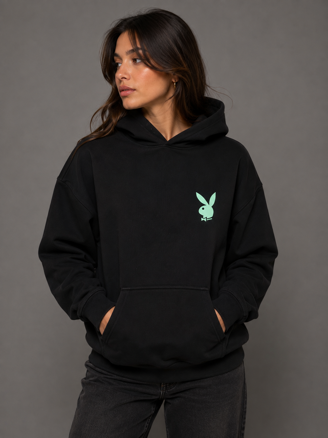 A woman wearing a black pullover hoodie with a small mint green Playboy bunny logo on the left chest, posed against a solid dark gray studio background. #Women