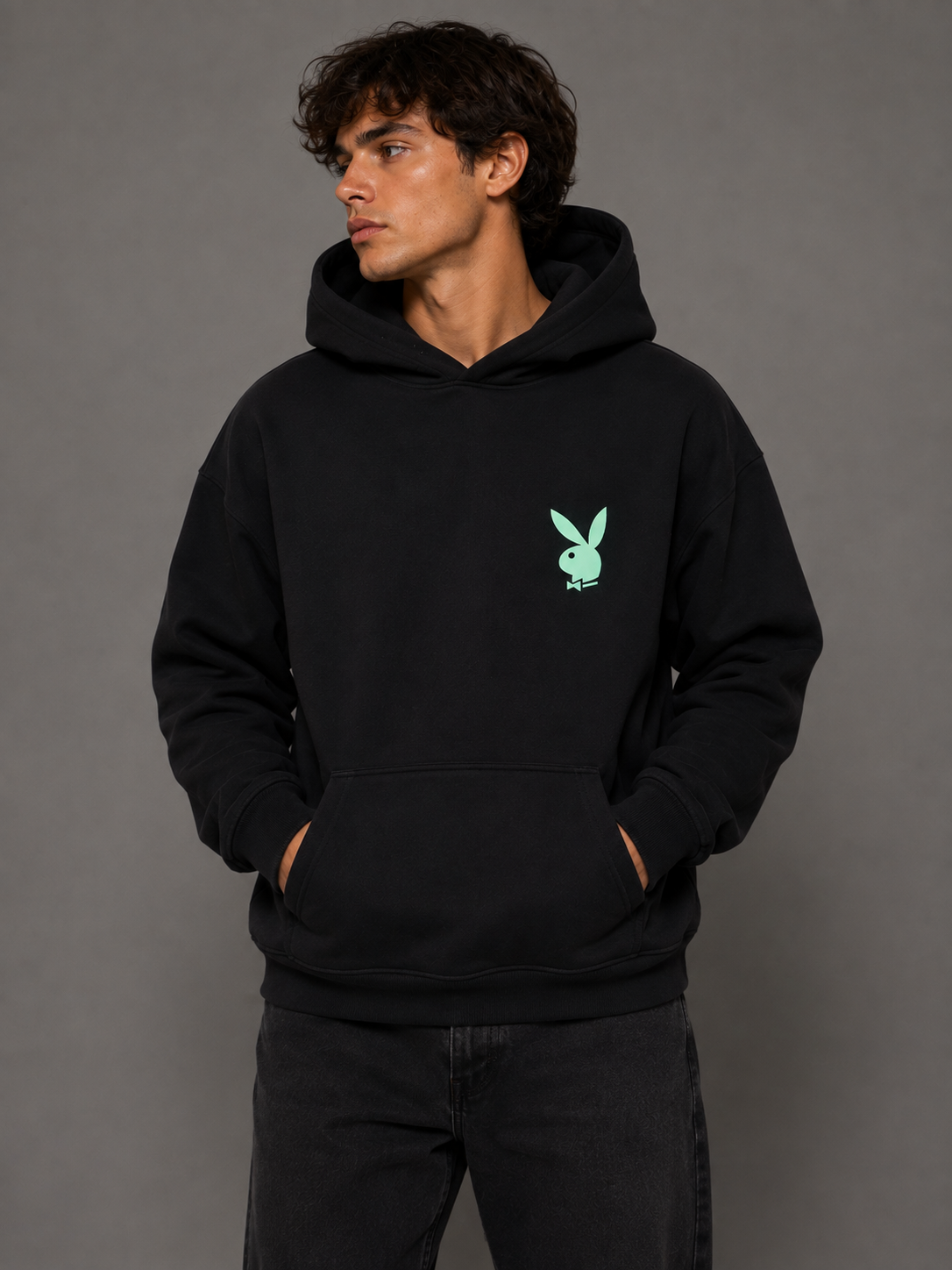A man wearing a black pullover hoodie with a small mint green Playboy bunny logo on the left chest, posed against a solid gray studio background.