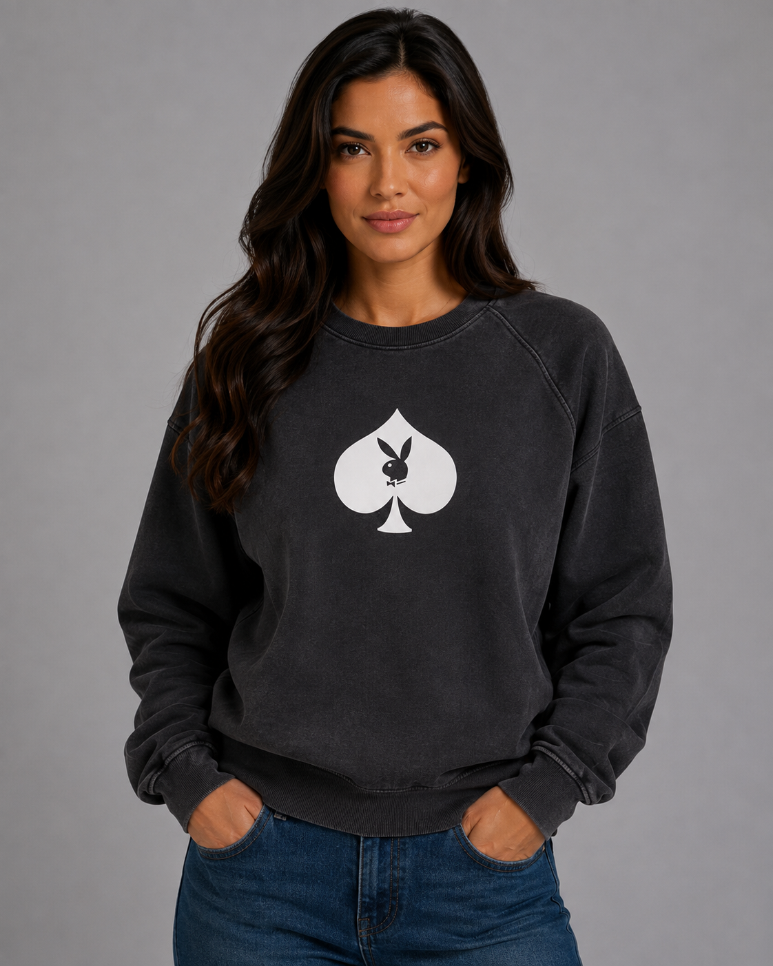 A woman wearing a dark charcoal gray sweatshirt with a white Playboy spade graphic on the chest, paired with blue jeans, against a solid gray studio background.#Women